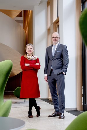  Ein Mann und eine Frau, beide in professioneller Kleidung, stehen lächelnd nebeneinander vor einem modernen Bürogebäude.