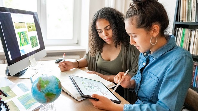 Zwei junge Frauen sitzen an einem Computer und Tablet und sprechen über Klimaschutzmaßnahmen.